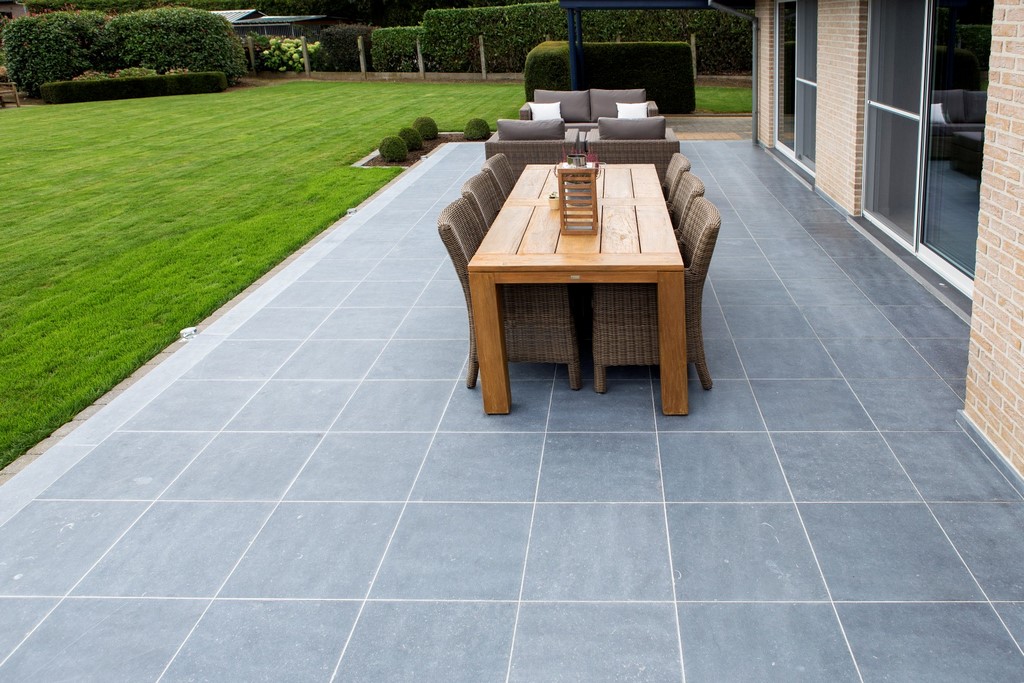 Een terras uit blauwe steen waarop een grote, houten tuintafel staat en een loungeset.