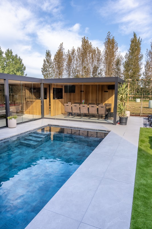 Een tuin met poolhouse, buitenterras en een zwembadrand in keramische tegels. 