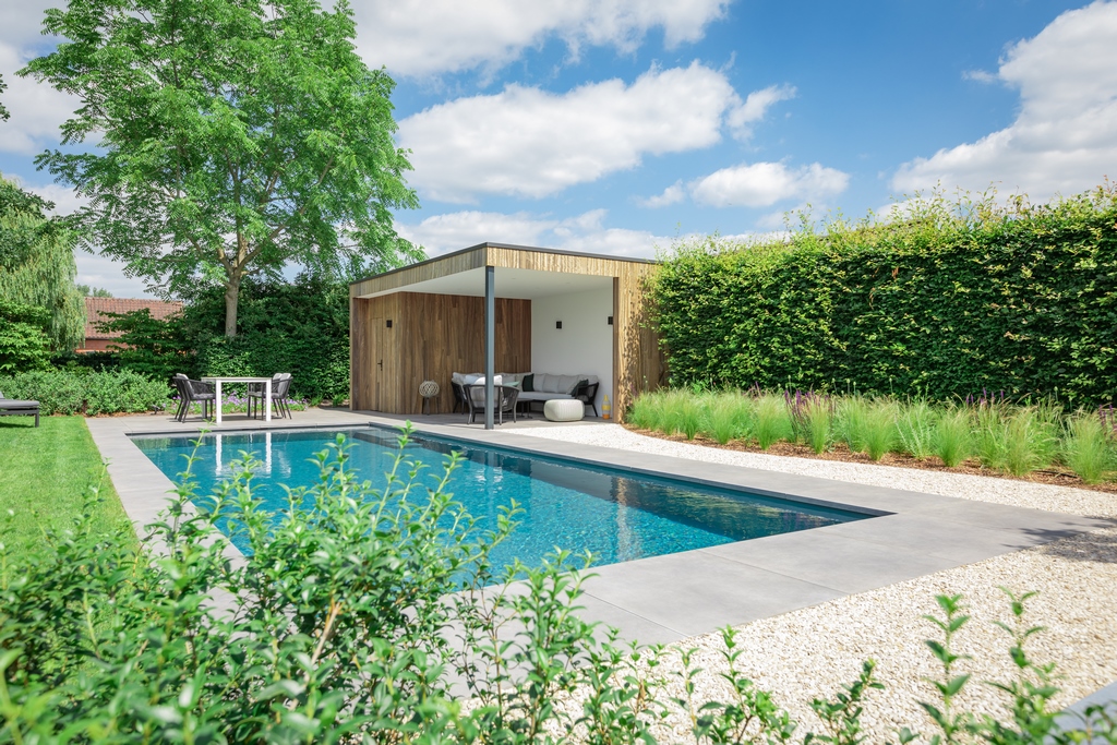 Een groene tuin met rechthoekig zwembad met zwembadranden in natuursteen en een poolhouse.